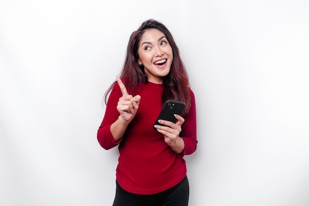 Donna asiatica eccitata che indossa la parte superiore rossa che punta allo spazio della copia sopra di lei mentre tiene il telefono isolato da uno sfondo bianco