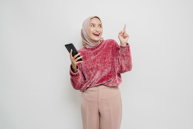 Donna asiatica eccitata che indossa un maglione rosa e hijab che punta allo spazio della copia sopra di lei mentre tiene il telefono isolato da uno sfondo bianco