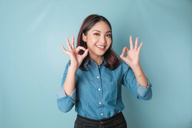 Donna asiatica eccitata che indossa una camicia blu che dà un gesto della mano OK isolato da uno sfondo blu