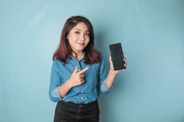 Donna asiatica eccitata che indossa una camicia blu che punta allo spazio di copia sul suo smartphone isolato da sfondo blu