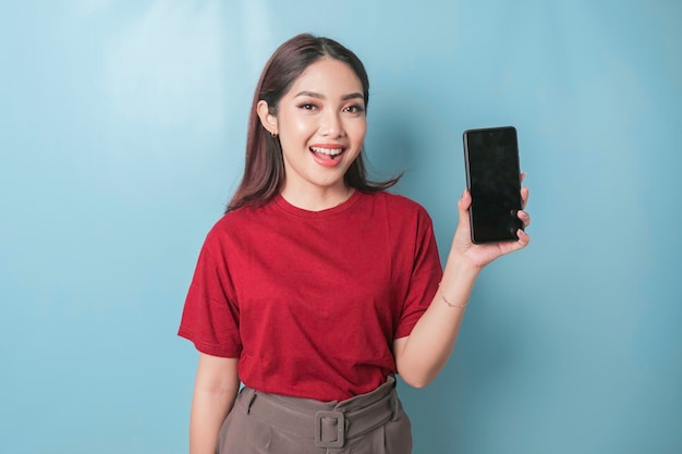Donna asiatica eccitata che indossa una maglietta rossa che mostra e indica lo spazio della copia sul suo smartphone isolato da sfondo blu