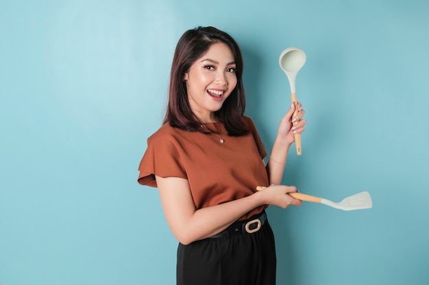 Donna asiatica eccitata che tiene articoli da cucina e sorridente isolati da sfondo blu