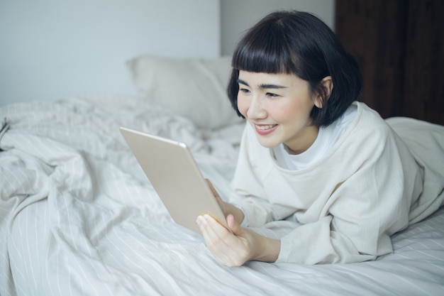 Donna asiatica felice che usando taplet per lavoro alla camera da letto.