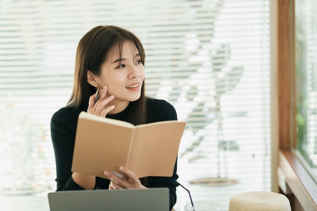 Donna asiatica freelance che sorride usando la penna che fa gli avvisi il suo taccuino e lavora al computer portatile sul tavolo a casa Imprenditrice che lavora per la sua attività al bar Lavoro d'affari al concetto di caffetteria