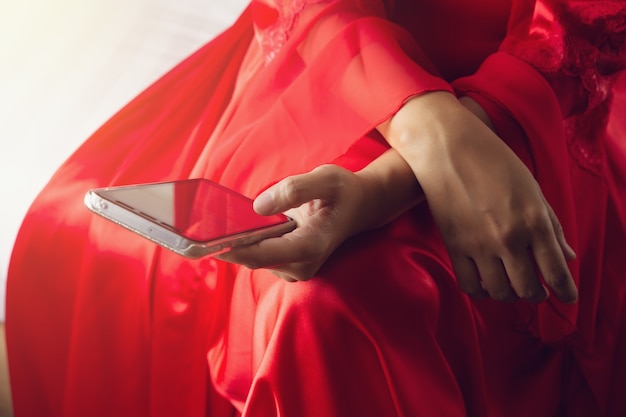 Donna asiatica in abiti di raso rosso utilizzando un telefono cellulare mentre era seduto su un letto di notte