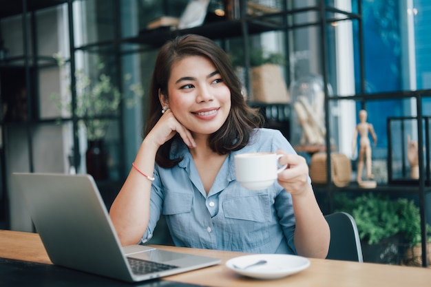 Donna asiatica in camicia blu nel caffè bevente del caffè e nell'uso della commercializzazione online di affari di lavoro del computer portatile