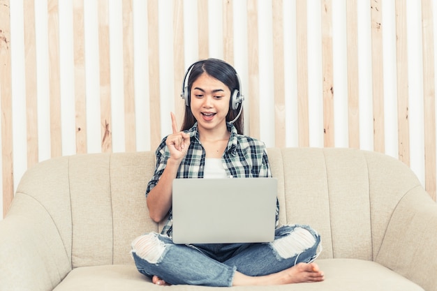 Donna asiatica in jeans e camicia a quadri. Ritratto di una giovane donna felice seduta sul divano e che lavora o studia online