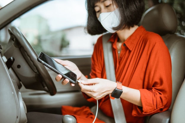 Donna asiatica in maschera protettiva alla guida di un'auto su strada. caricare il suo telefono