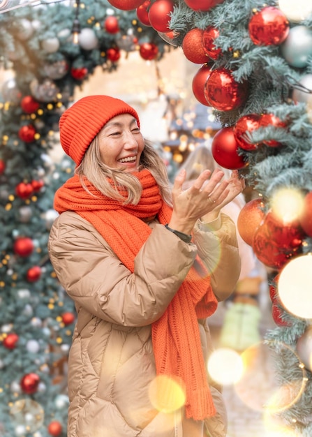 Donna asiatica in sciarpa arancione e cappello che cammina nel mercatino di Natale decorato con luci natalizie