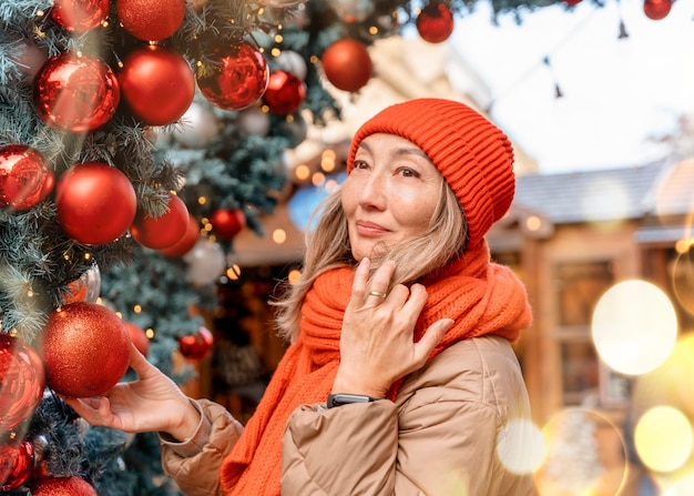 Donna asiatica in sciarpa arancione e cappello che cammina nel mercatino di Natale decorato con luci natalizie