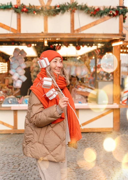 Donna asiatica in sciarpa arancione e cappello che cammina nel mercatino di Natale decorato con luci natalizie