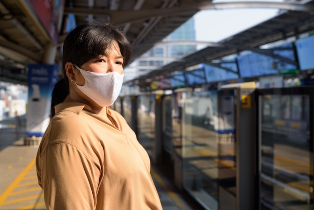 Donna asiatica in sovrappeso con maschera che pensa e in attesa alla stazione del treno di alianti