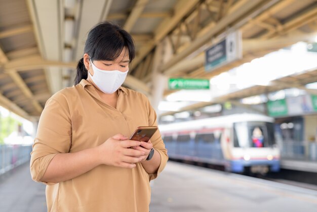 Donna asiatica in sovrappeso con maschera utilizzando il telefono alla stazione del treno di alianti