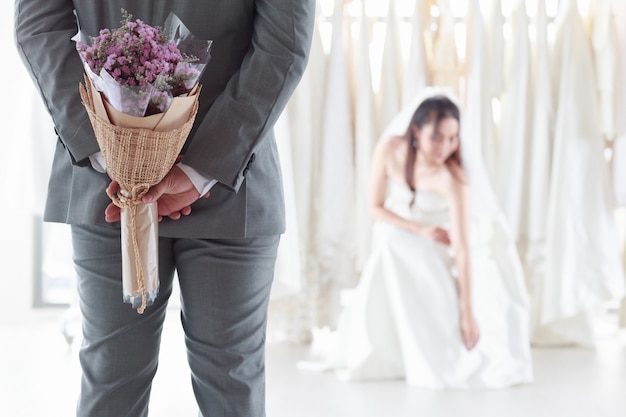 Donna asiatica in un abito da sposa Sposo sorridente. Lo sposo che indossa un abito grigio tiene in mano un mazzo di fiori viola per sorprendere la sposa che siede nel camerino. Il giorno migliore del matrimonio di concetto.