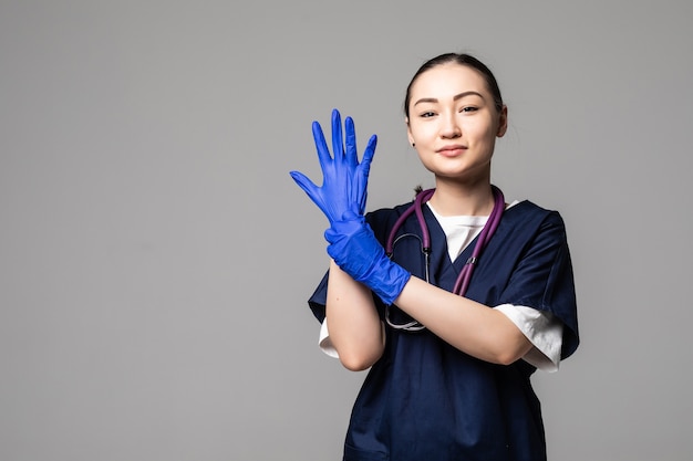 Donna asiatica in uniforme medica e maschera facciale che indossa guanti protettivi isolati su muro bianco