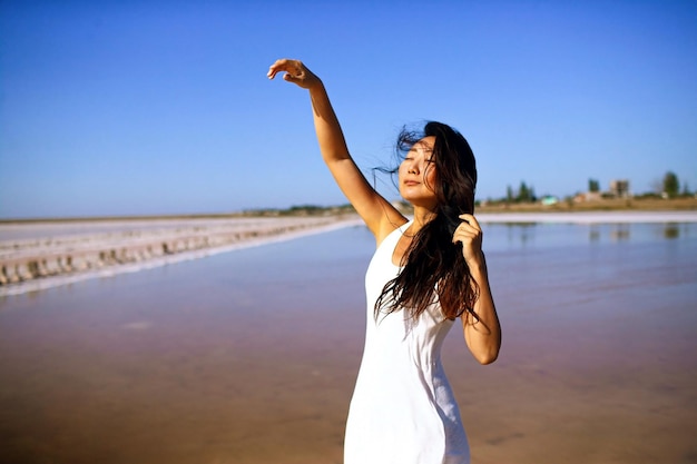 Donna asiatica in vestito bianco con lunghi capelli scuri che danza nel vento sulla spiaggia del mare rosa.