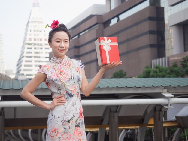 Donna asiatica in vestito cinese che tiene il contenitore di regalo rosso a disposizione con il fondo della città