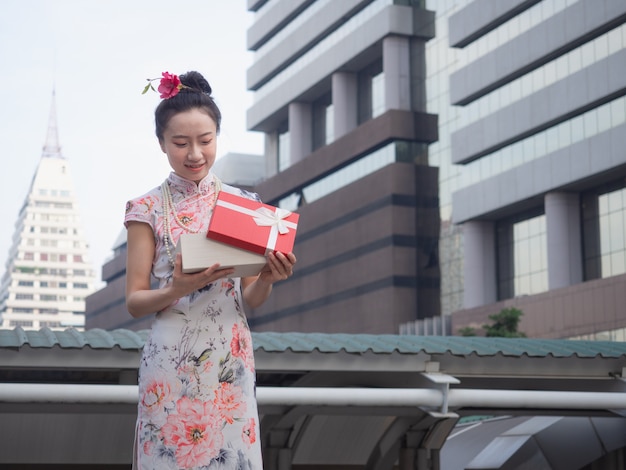 Donna asiatica in vestito cinese che tiene il contenitore di regalo rosso a disposizione con il fondo della città