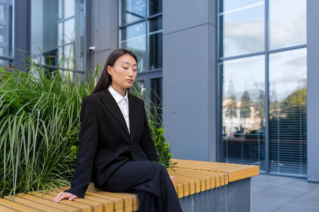 Donna asiatica licenziata triste che si siede sulla panchina depressa vicino all'ufficio