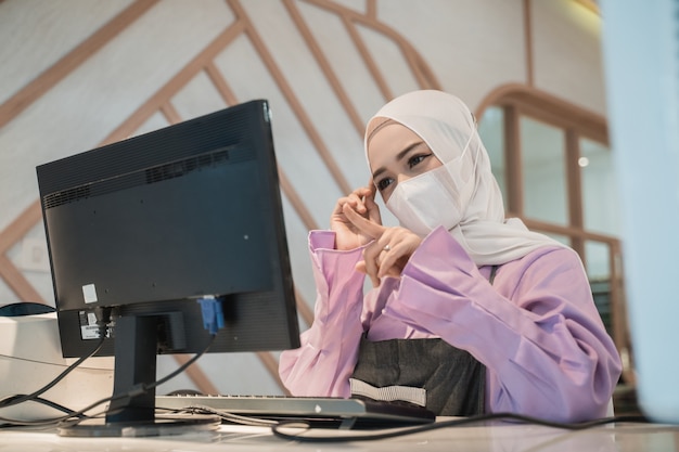 Donna asiatica musulmana che lavora utilizzando il pc mentre indossa la maschera medica per la protezione in ufficio