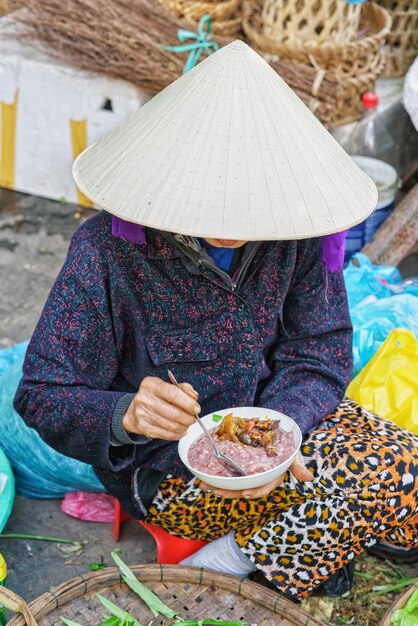 Donna asiatica nel tradizionale cappello vietnamita che mangia cibo nel mercato di strada a Hoi An, Vietnam. Senza faccia