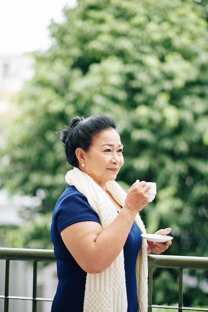 Donna asiatica pensierosa seria in piedi sul balcone e beve caffè mattutino