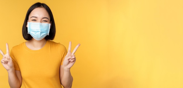 Donna asiatica positiva sorridente che indossa una maschera medica da covid durante la pandemia che mostra la pace vsig...