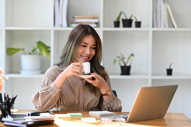 Donna asiatica premurosa di affari che beve caffè caldo e legge la posta elettronica sul computer portatile