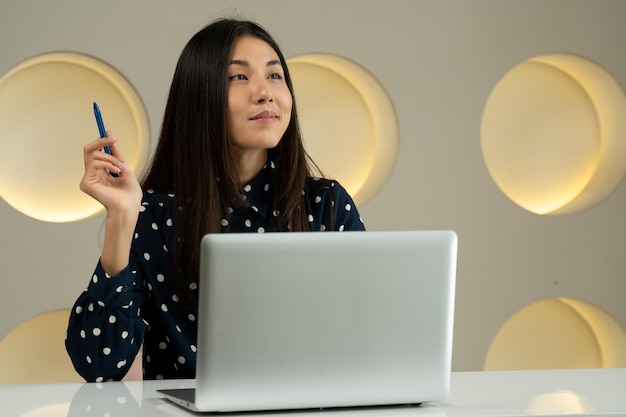 Donna asiatica premurosa di affari che lavora ad un computer che sorride e che esamina il pensiero di distanza