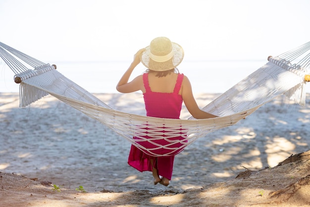 Donna asiatica rilassante nell'amaca sulla spiaggia tropicale goditi la sua libertà e aria fresca indossando cappello e vestiti eleganti Felice turista sorridente ai tropici in vacanza di viaggio