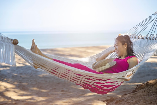 Donna asiatica rilassante nell'amaca sulla spiaggia tropicale goditi la sua libertà e aria fresca indossando cappello e vestiti eleganti Felice turista sorridente ai tropici in vacanza di viaggio