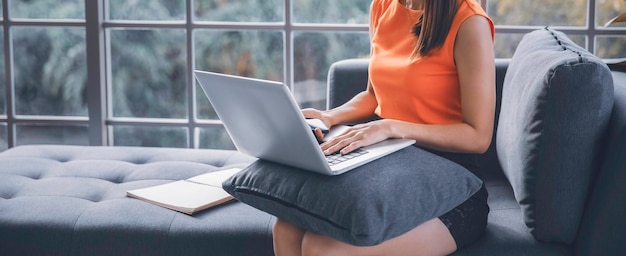 Donna asiatica seduta sul divano utilizzando smartphone e computer portatile che lavora a casa, concetto di lavoro mobile con spazio di copia