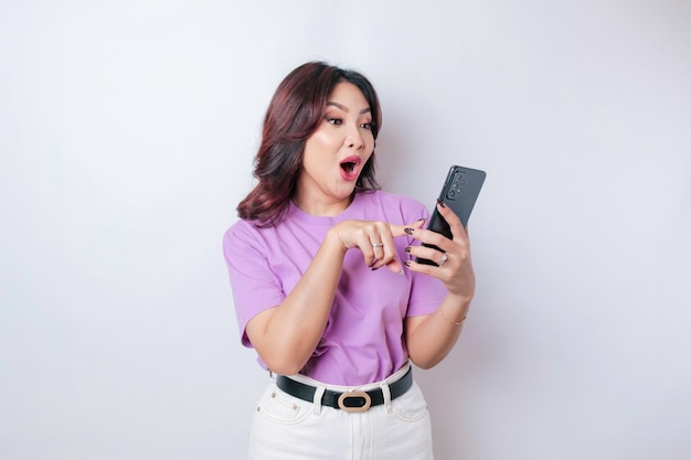 Donna asiatica sorpresa che indossa una maglietta viola lilla che punta al suo smartphone isolato da sfondo bianco