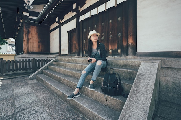 Donna asiatica sorridente con il cappello che prende la macchina fotografica professionale nelle mani che si siedono sulle scale fuori. viaggiatore della ragazza che riposa all'aperto dopo la visita al tempio di shitennoji con lo zaino accanto. femmina che guarda da parte.