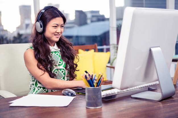 Donna asiatica sorridente con le cuffie che utilizza computer nell&#39;ufficio