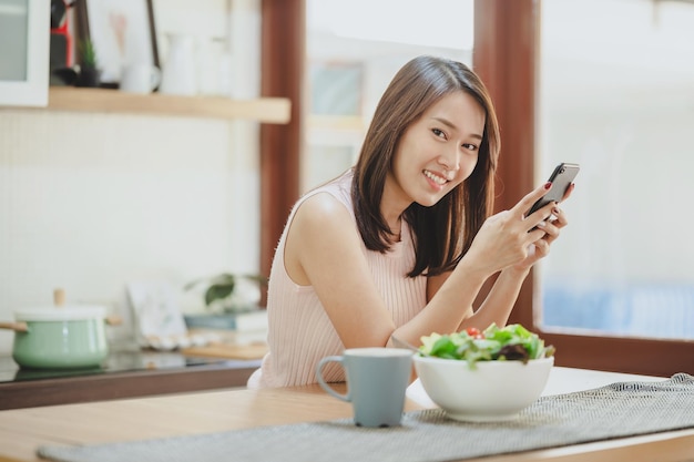 Donna asiatica sorridente felice che utilizza il cellulare dello smartphone a casa in cucina con un'insalatiera sul tavolo