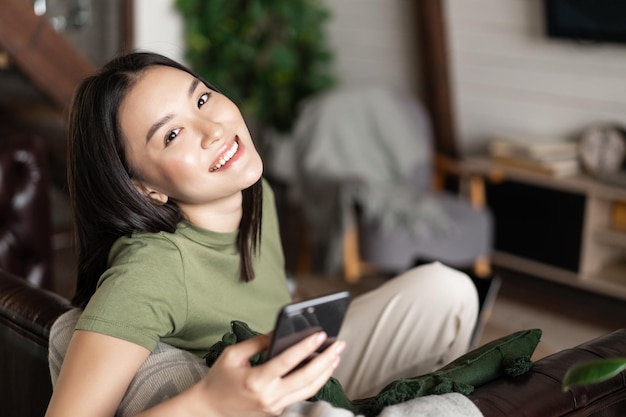 Donna asiatica sorridente sognante che utilizza il telefono cellulare seduta e rilassante a casa