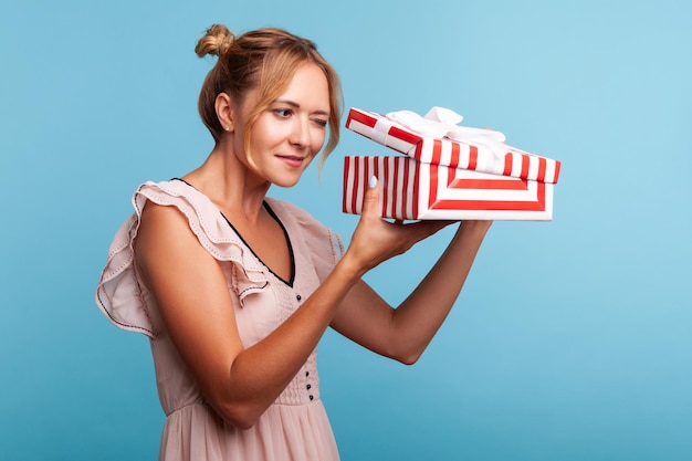 Donna astuta con capelli biondi e due pacchi unboxing confezione regalo a strisce rosse, guardando all'interno con curiosa espressione facciale, regalo di compleanno. Studio indoor girato isolato su sfondo blu.