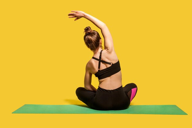 Donna atleta vista posteriore con crocchia per capelli in cima attillata e pantaloni seduta nella posizione del loto sul tappetino da palestra praticando yoga, piegandosi di lato con la mano sollevata, facendo allenamenti di stretching. girato in studio isolato