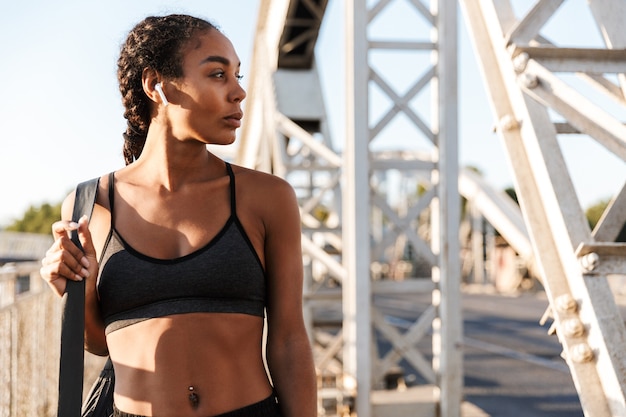 donna atletica afroamericana in abbigliamento sportivo utilizzando auricolari mentre si cammina con la borsa sul vecchio ponte