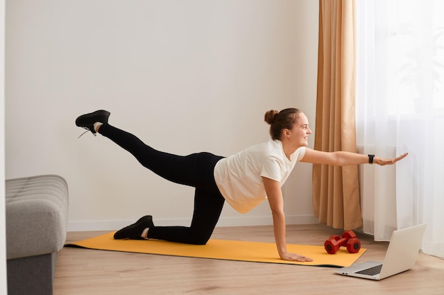 Donna atletica caucasica che indossa abbigliamento sportivo in piedi nella posa della tavola con la gamba del braccio sollevata, facendo allenamento yoga fitness a casa, allungando l'esercizio di base sul tappetino sul pavimento.