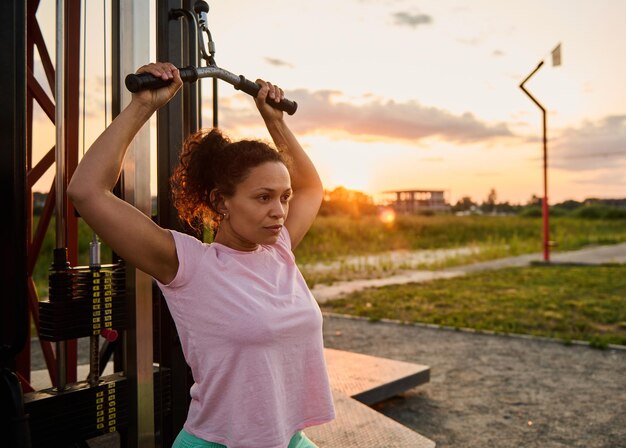 Donna atletica che fa esercizi per le braccia utilizzando il simulatore stazionario in un parco contro il tramonto