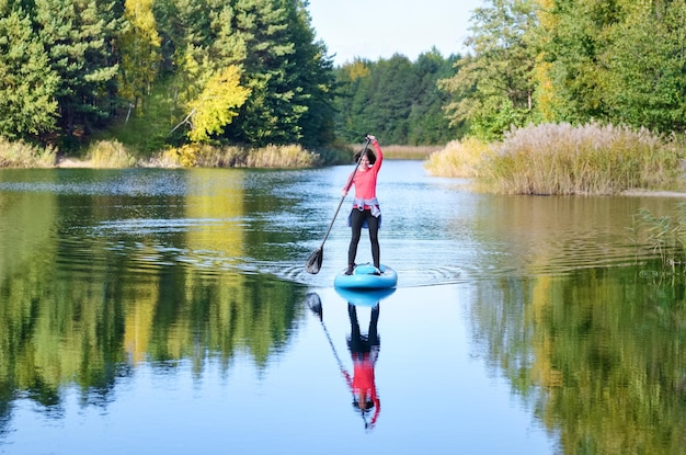 Donna attiva che pagaia tavola SUP sul bellissimo lago, paesaggio forestale autunnale e natura sullo sfondo