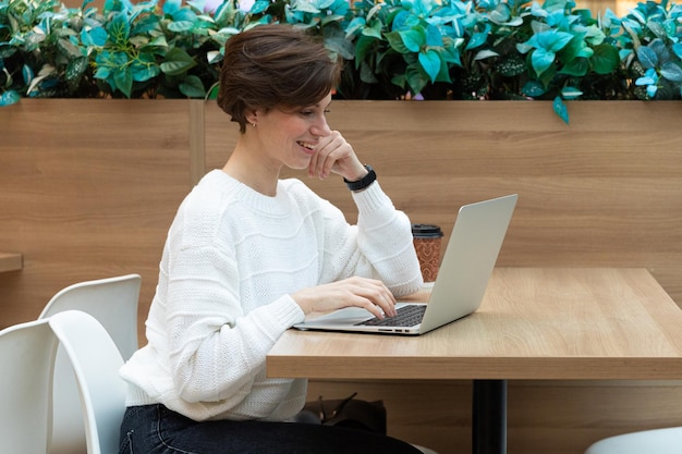 Donna attraente adulta che si siede ad un centro commerciale ad un tavolo e che lavora ad un computer portatile