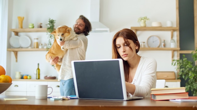 Donna attraente che cerca di lavorare da remoto mentre suo marito tiene tra le braccia un cane e balla