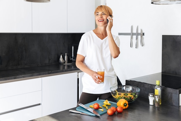 Donna attraente che cucina il pranzo mentre si trovava in cucina a casa, parlando al telefono cellulare