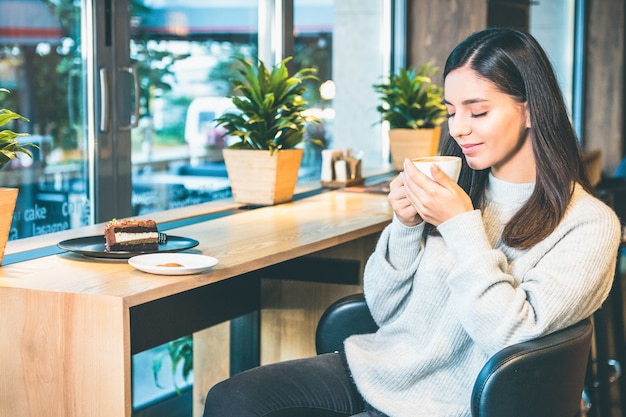 Donna attraente che giudica una tazza di caffè elegante con la seduta chiusa degli occhi dalla finestra