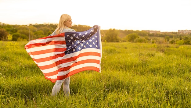 donna attraente che tiene una bandiera americana nel vento in un campo. Paesaggio estivo contro il cielo blu. Orientamento orizzontale.