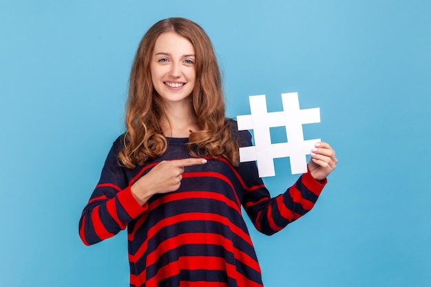 Donna attraente con capelli mossi in maglione che punta a hashtag bianco, post sui social media alla moda e blog, contenuti web virali, promozione su Internet. Studio indoor girato isolato su sfondo blu.