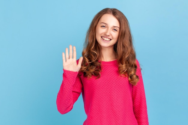 Donna attraente con capelli ondulati che indossa un pullover rosa con il braccio alzato che dice ciao guardando la fotocamera con un'espressione felice e un sorriso a trentadue denti Colpo in studio al coperto isolato su sfondo blu